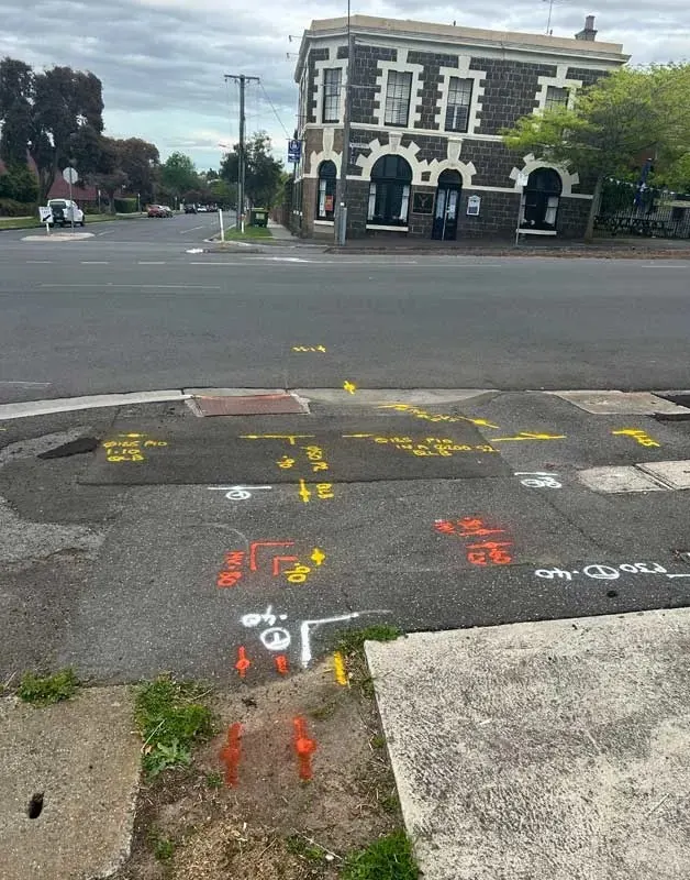 Utility mark-out in yellow, orange and white paint on a footpath and kerb, with service annotations including