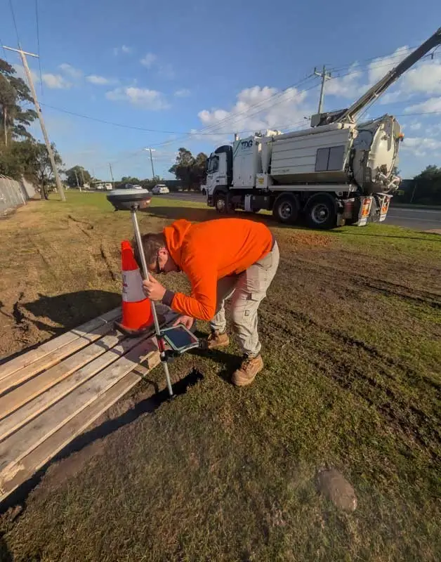 A Geoscan surveyor in hi-vis holding a GNSS rover pole in an excavation hole, with a vacuum excavation truck behind.